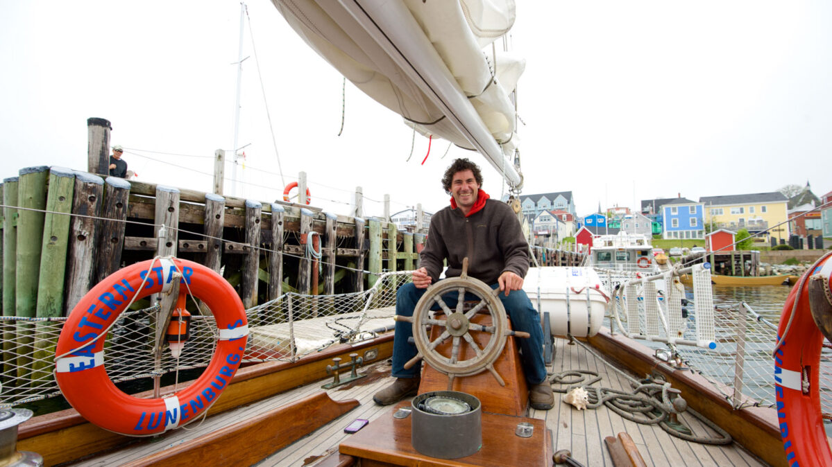 A man sitting on a boat holding a stirring wheel.