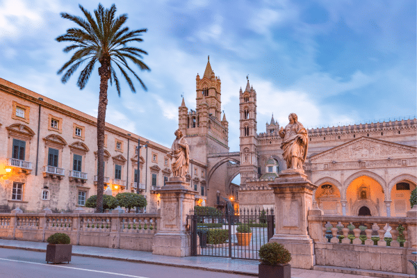 Image of the Norman Cathedral taken from outside the front gates. Featuring ornate statues and spires, along with a tall, lush palm tree