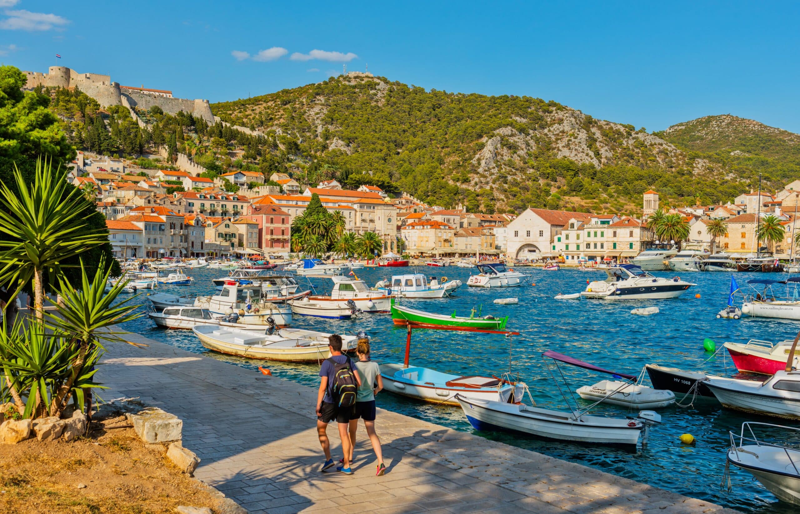 Two travelers walking along Hvar's waterfront in Croatia, overlooking colorful stone buildings, fishing boats, the Adriatic Sea, and the hilltop fortress.