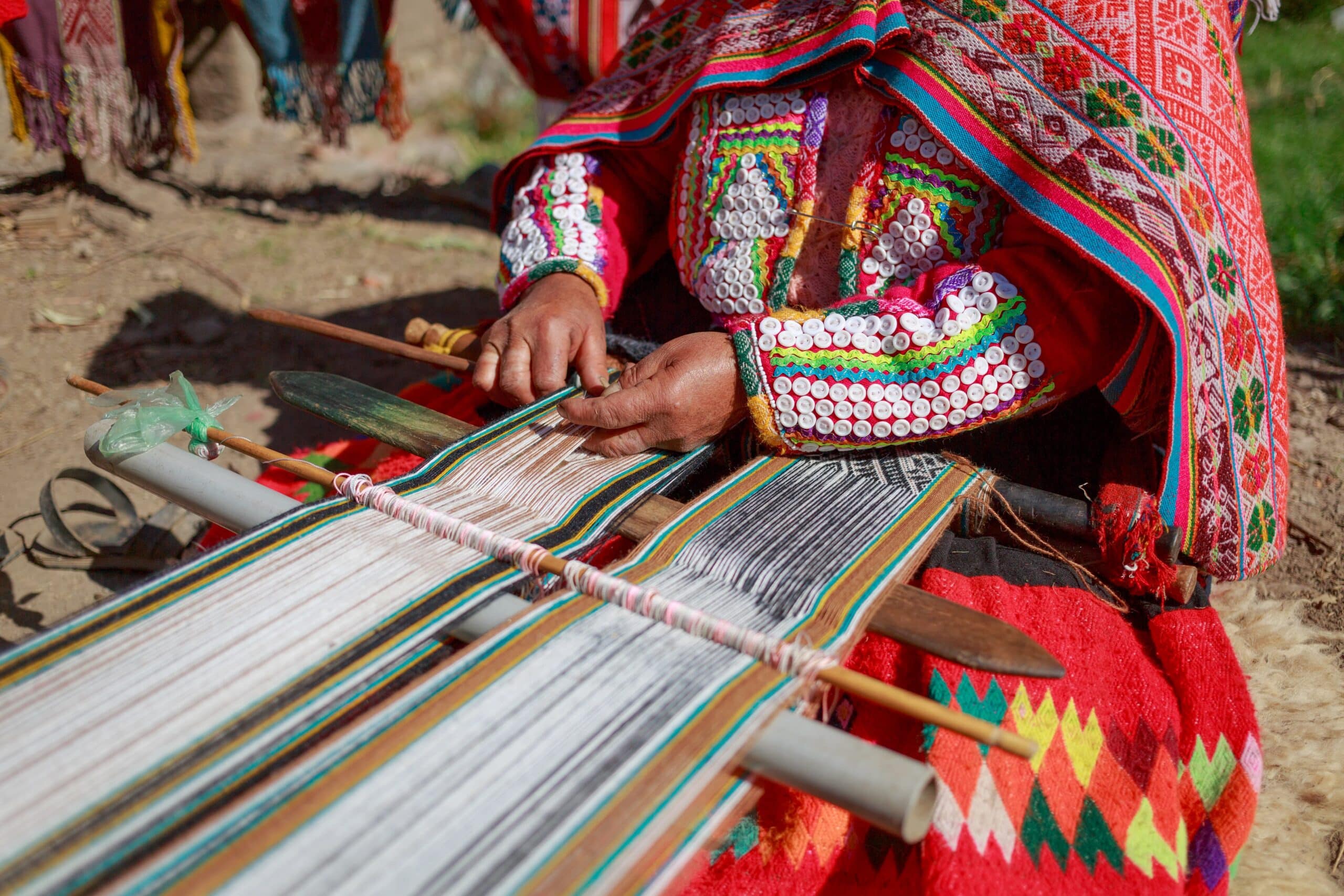 Quechua textile artisan in colorful traditional Andean clothing weaving a striped textile on a loom in Huilloc, Peru's Sacred Valley.